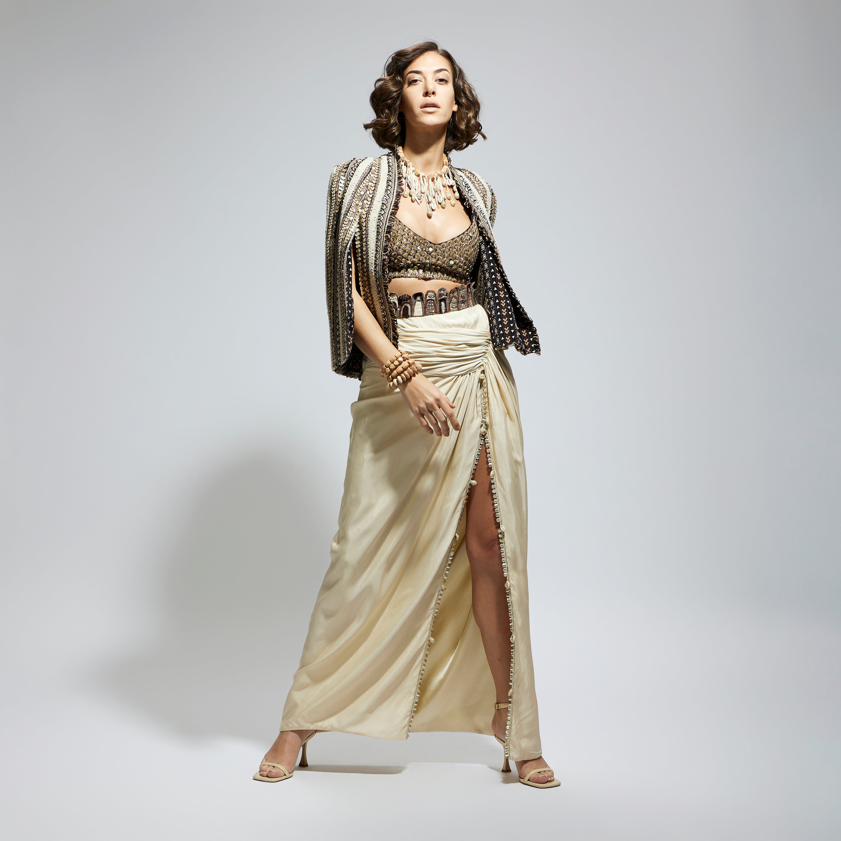 SAMSARA: EMBELLISHED & TEXTURED CAPE JACKET PAIRED WITH METALLIC SCALLOP BUSTIER AND IVORY HIGH SLIT SKIRT